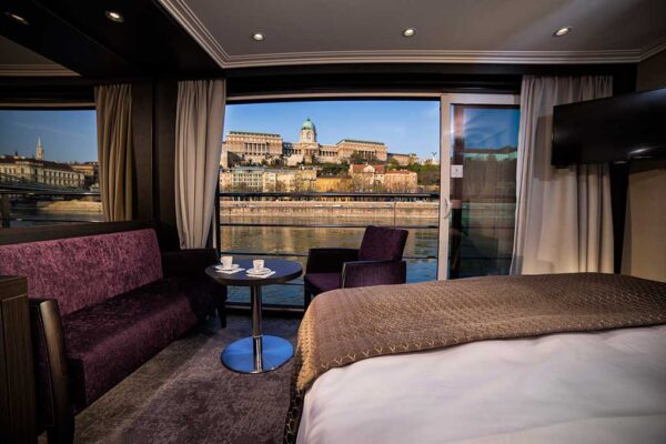 Panorama Suite view of buildings, purple couch with table and chair, bed and TV - Avalon Waterways